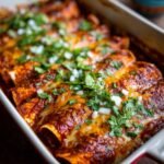 Close-up of Birria Enchiladas in a baking dish, topped with cheese, cilantro, and diced onions.