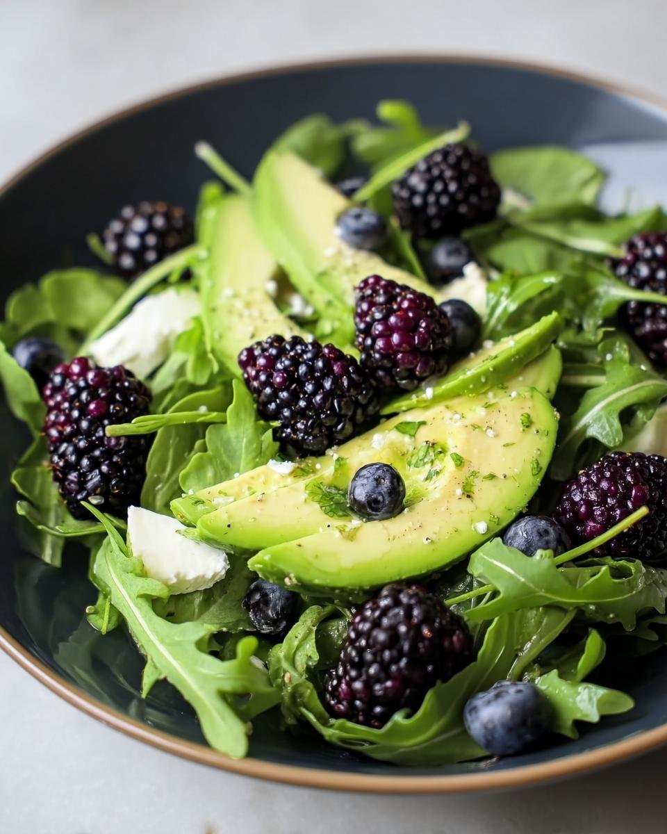 A vibrant bowl of Blackberry, Avocado & Arugula Salad with blueberries and cheese.