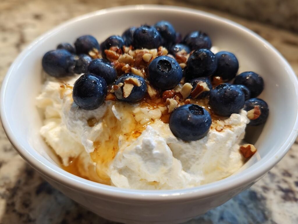 A bowl of Blueberry Cottage Cheese Breakfast with fresh blueberries, chopped nuts, and a drizzle of honey.