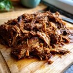 Close-up of caramelized pulled beef brisket on a wooden cutting board, glistening with sauce.