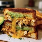 Stacked Chicken Avocado Melt Sandwich with melted cheese and toasted bread, served with fries.