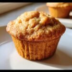 A close-up of a golden brown Cottage Cheese Muffin on a white plate, with another muffin blurred in the background.