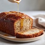 Maple-Bourbon Banana Bread loaf with a slice cut, drizzled with maple syrup on a plate.