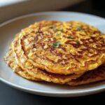 A stack of golden brown Red Lentil Pancakes on a white plate, garnished with fresh herbs.