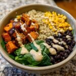 A vibrant Sweet Potato Buddha Bowl with roasted sweet potatoes, quinoa, corn, black beans, and spinach, drizzled with sauce.