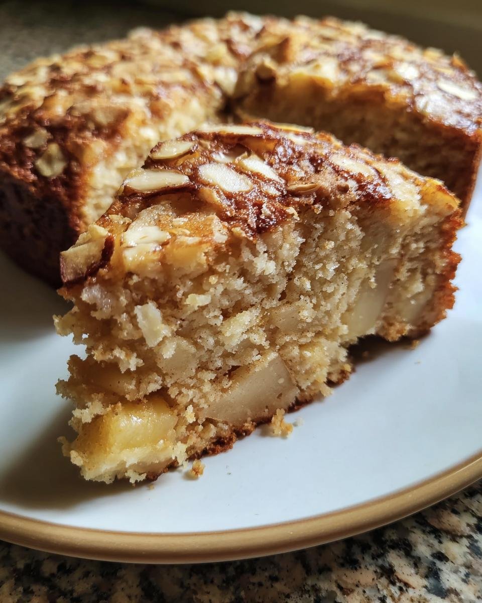Close-up of a slice of Oatmeal Apple Cake showing moist crumb and baked apple chunks, topped with toasted almonds.