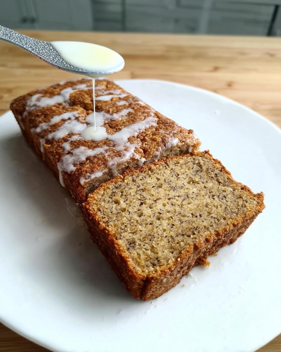 A slice of moist banana bread with a drizzle of glaze being poured from a spoon. Best Banana Bread Recipe with Ripe Bananas.