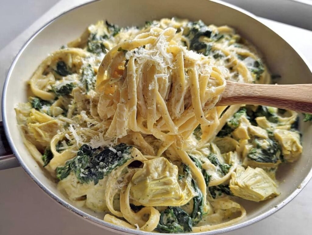 A wooden spoon lifts fettuccine coated in creamy spinach artichoke pasta sauce, topped with grated Parmesan.
