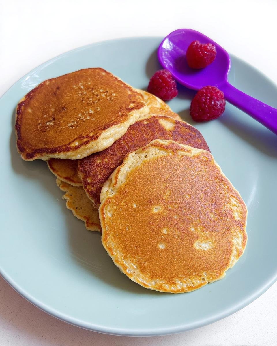 A stack of golden brown Three Ingredient Banana Pancakes served on a light blue plate with fresh raspberries and a purple spoon.